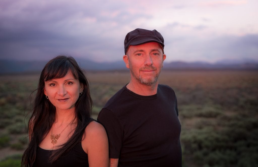 Two people, a woman and a man, face the camera smiling, with a prairie and hilly landscape in the background.
