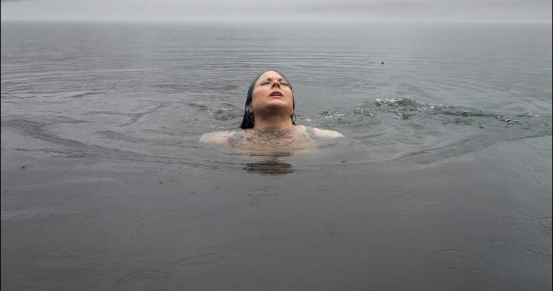 A woman swimming in water emerges from the surface with her eyes closed.