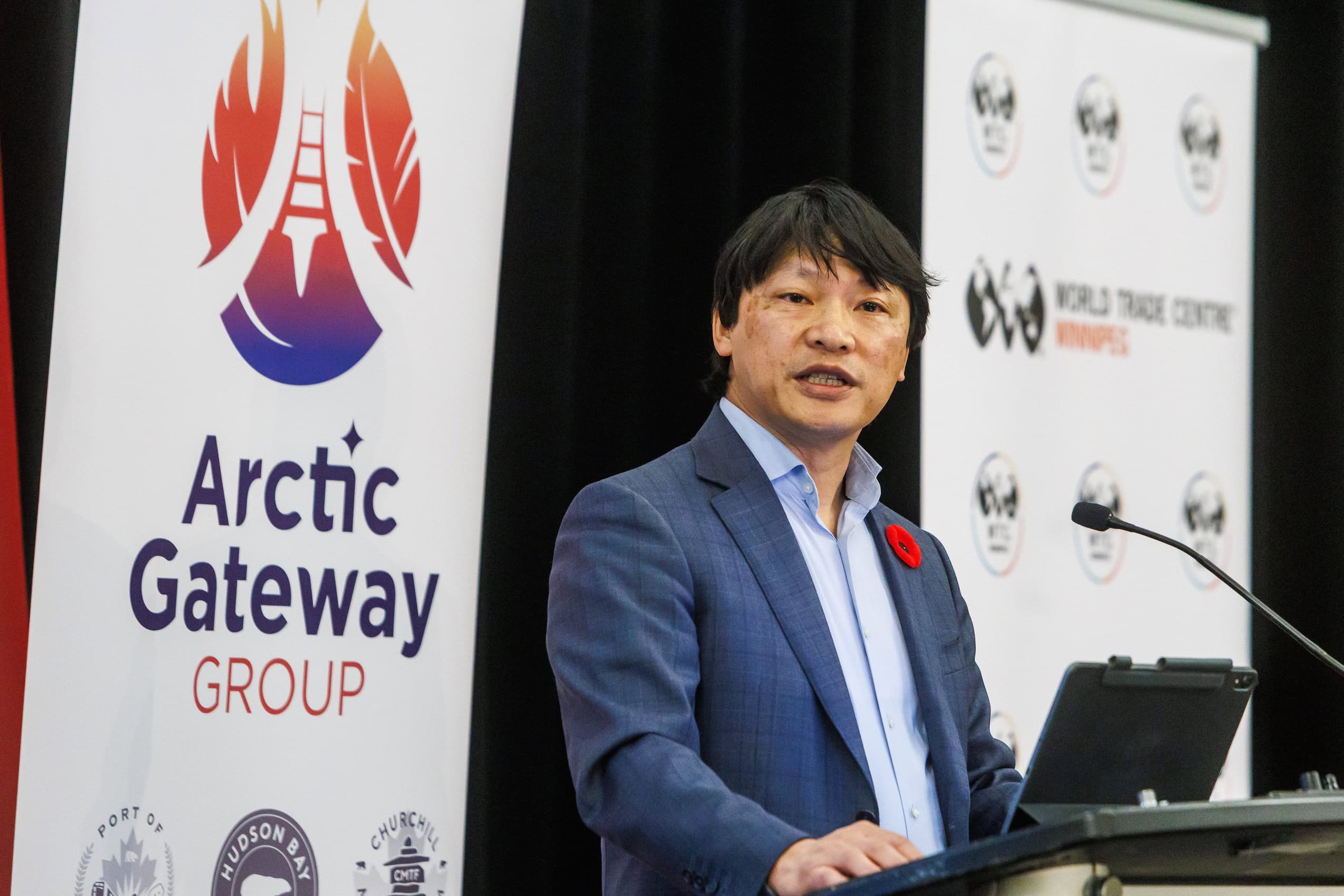 An Asian man with dark black hair speaks at a podium with an "Arctic Gateway Group" banner behind him.