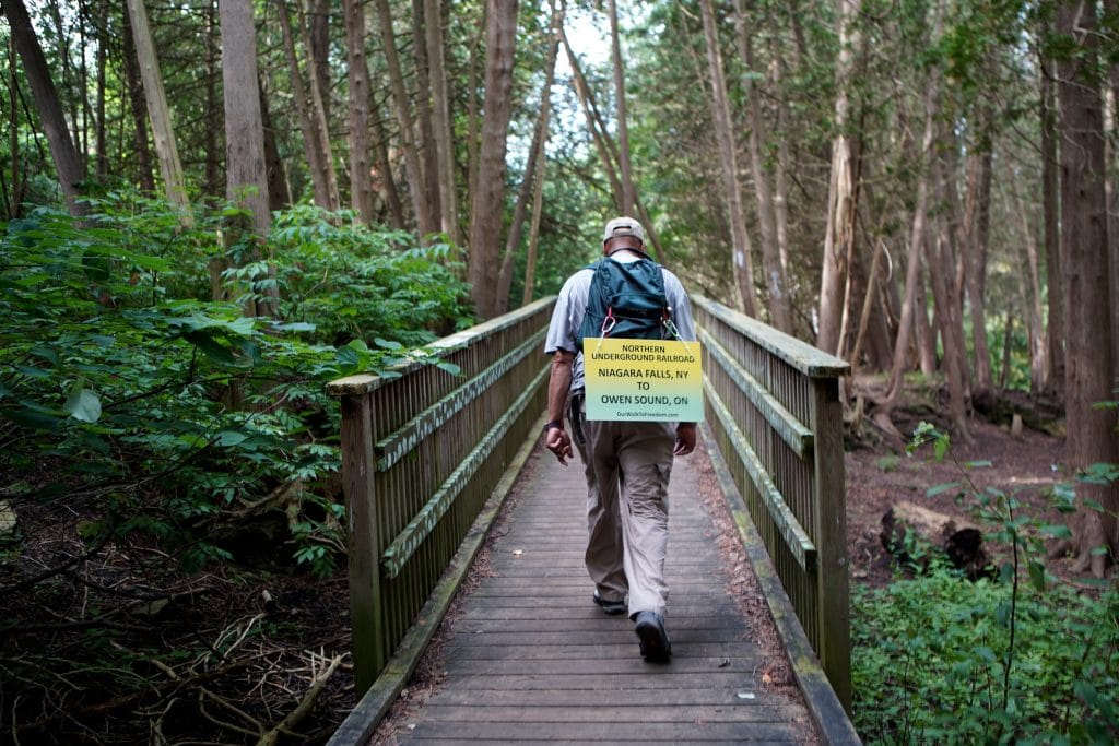 Ken Johnston walks through a park in Fergus, Ont., while retracing an Underground Railroad route from Niagara Falls to Owen Sound on his Walk to Freedom. He is seen from behind, with a sign that reads "Northern Underground Railroad, Niagara Falls, N.Y. to Owen Sound, ON."