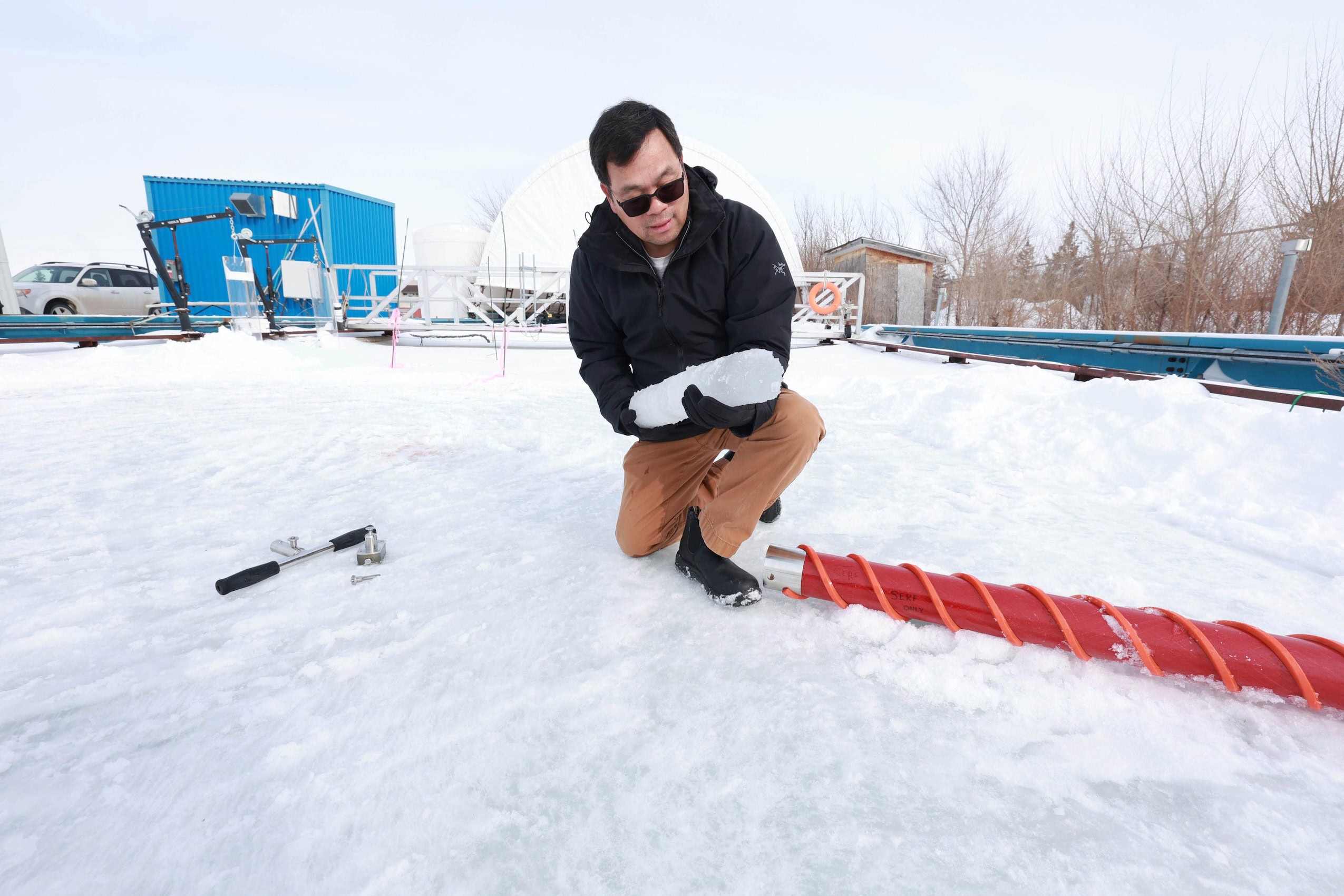 A man bends down over snow-covered ground, holding a large piece of ice in hand.