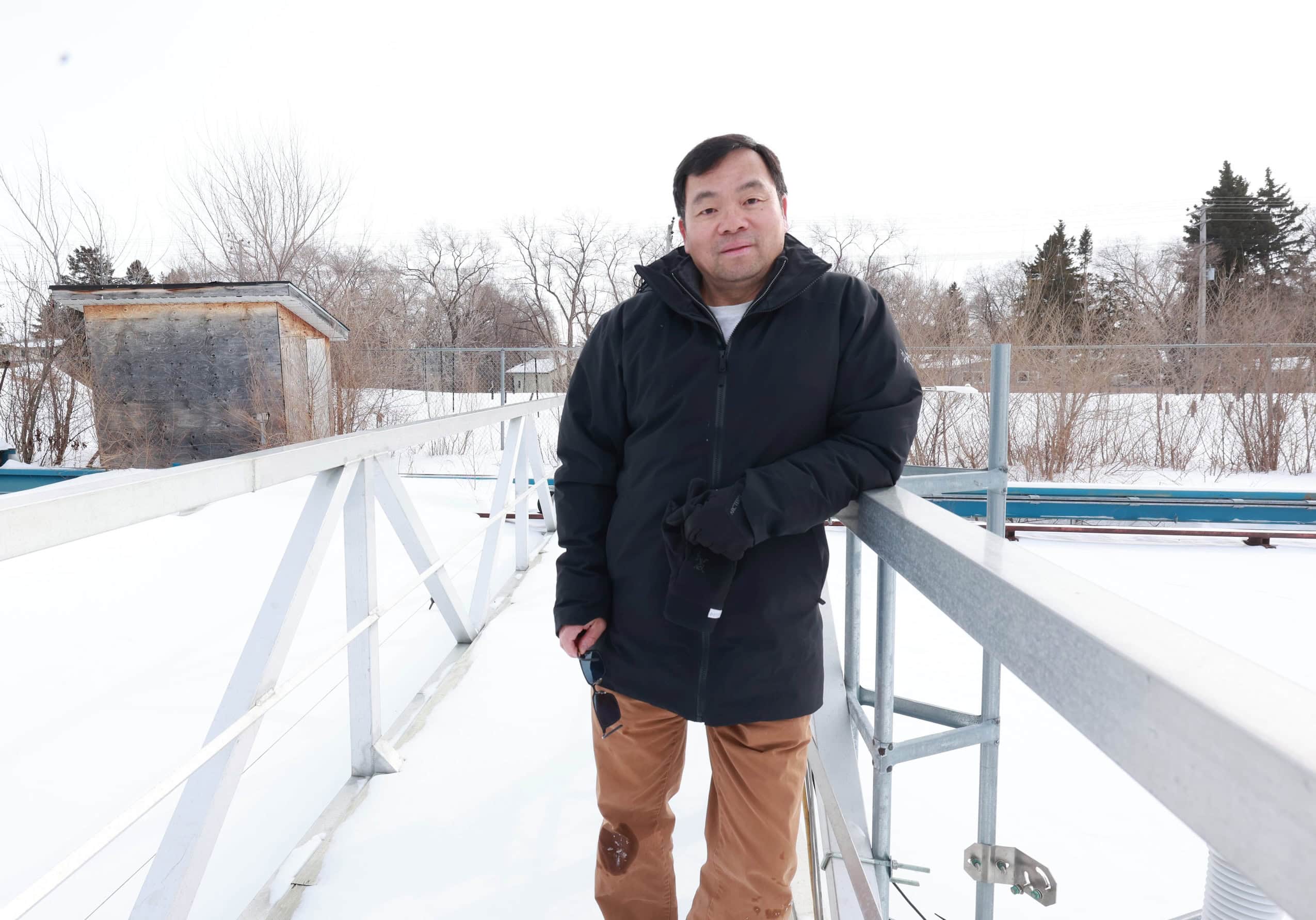 An Asian man leans against a bridge with a snow-covered landscape behind him.