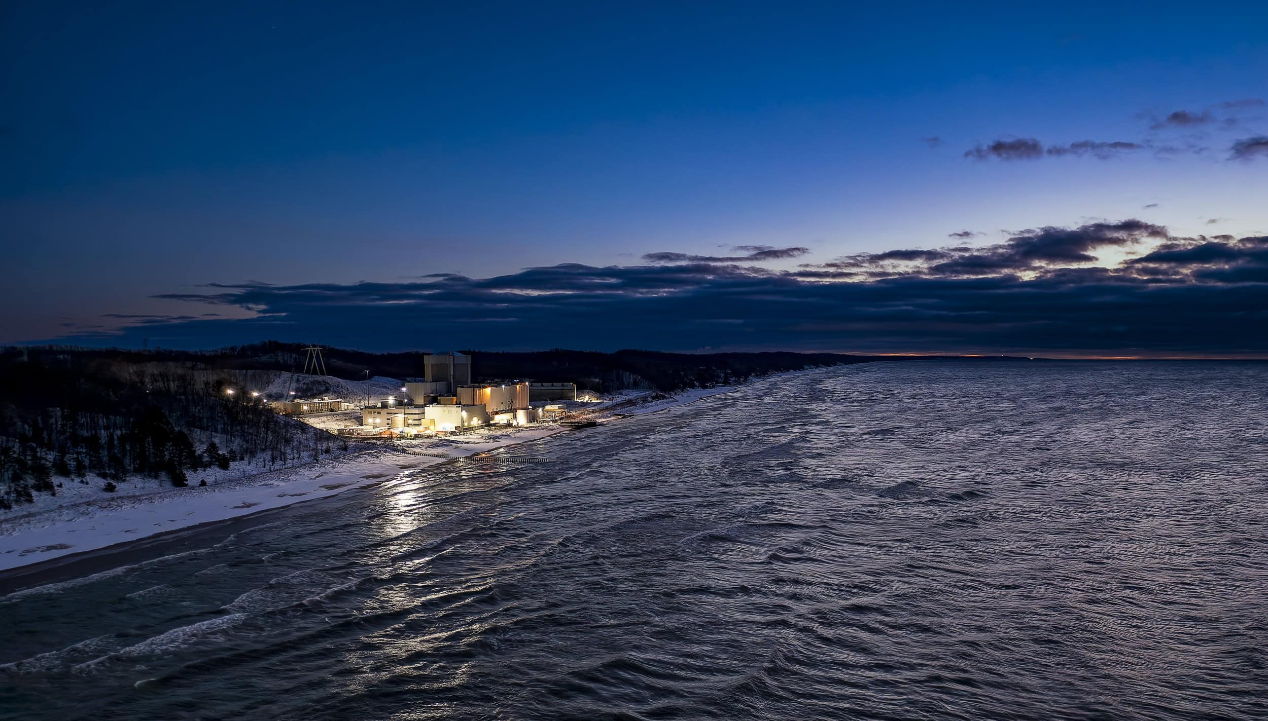 An industrial facility on the edge of a large lake as night falls.
