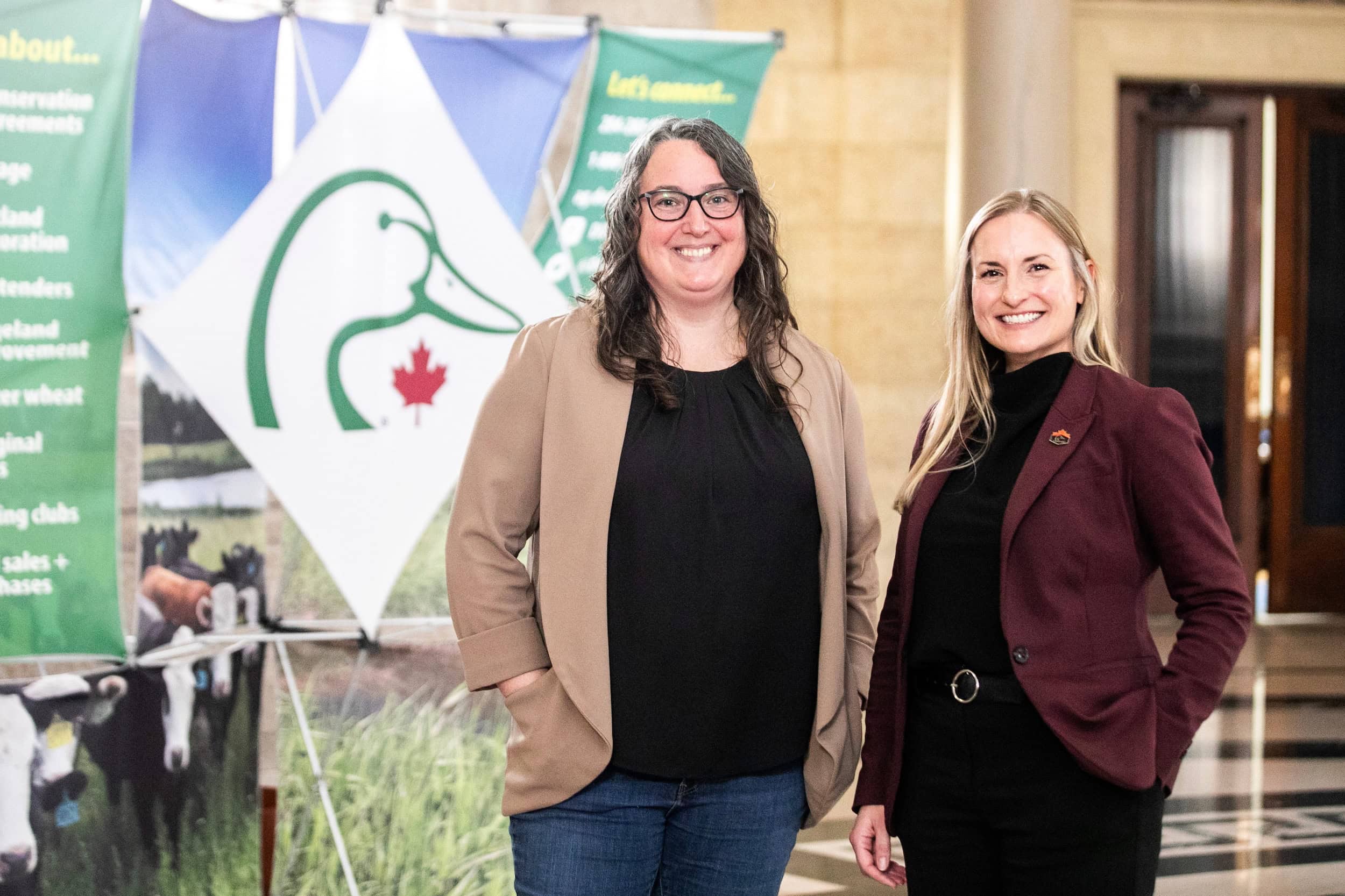 Two people pose indoors next to a Ducks Unlimited Canada sign