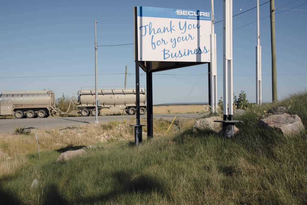 A blue and white sign that reads, "Thank you for your business" in blue cursive script. In the top right corner, white text on a blue bar reads "Secure Energy Services." The sign is mounted on three poles standing in the grass with a few small boulders around it. The sign is planted on a slop that rises toward the right of the frame. In the background, two tankers on a tanker truck are parked on the road