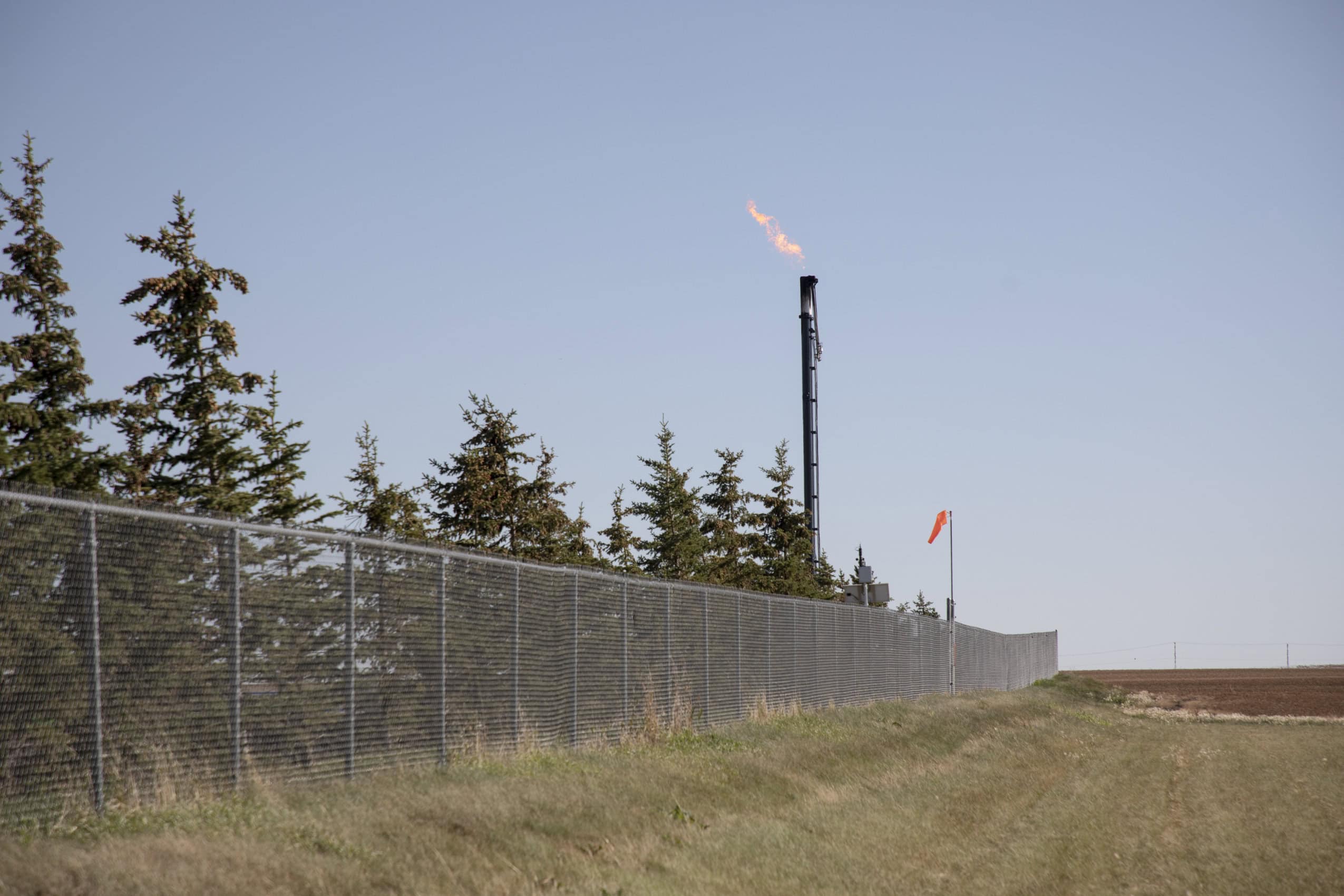 A lit flare stack stands behind a chain link fence and a row of small trees. There is a small orange windsock just beside the flare stack. The grass is cut short in the field on the other side of the fence. It's a sunny, clear day
