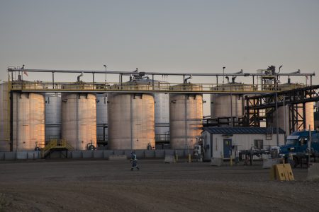 Nine large upright tanks stand along one edge of an oil and gas waste disposal facility. The sun is setting, casting a pink glow across the sides of the tanks. There's a metal walkway along with tops of the tanks. A working in a blue jump suit with reflective sites is walking across the gravel lot in front of the tanks. The blue cab of a parked heavy truck can be seen in the right corner