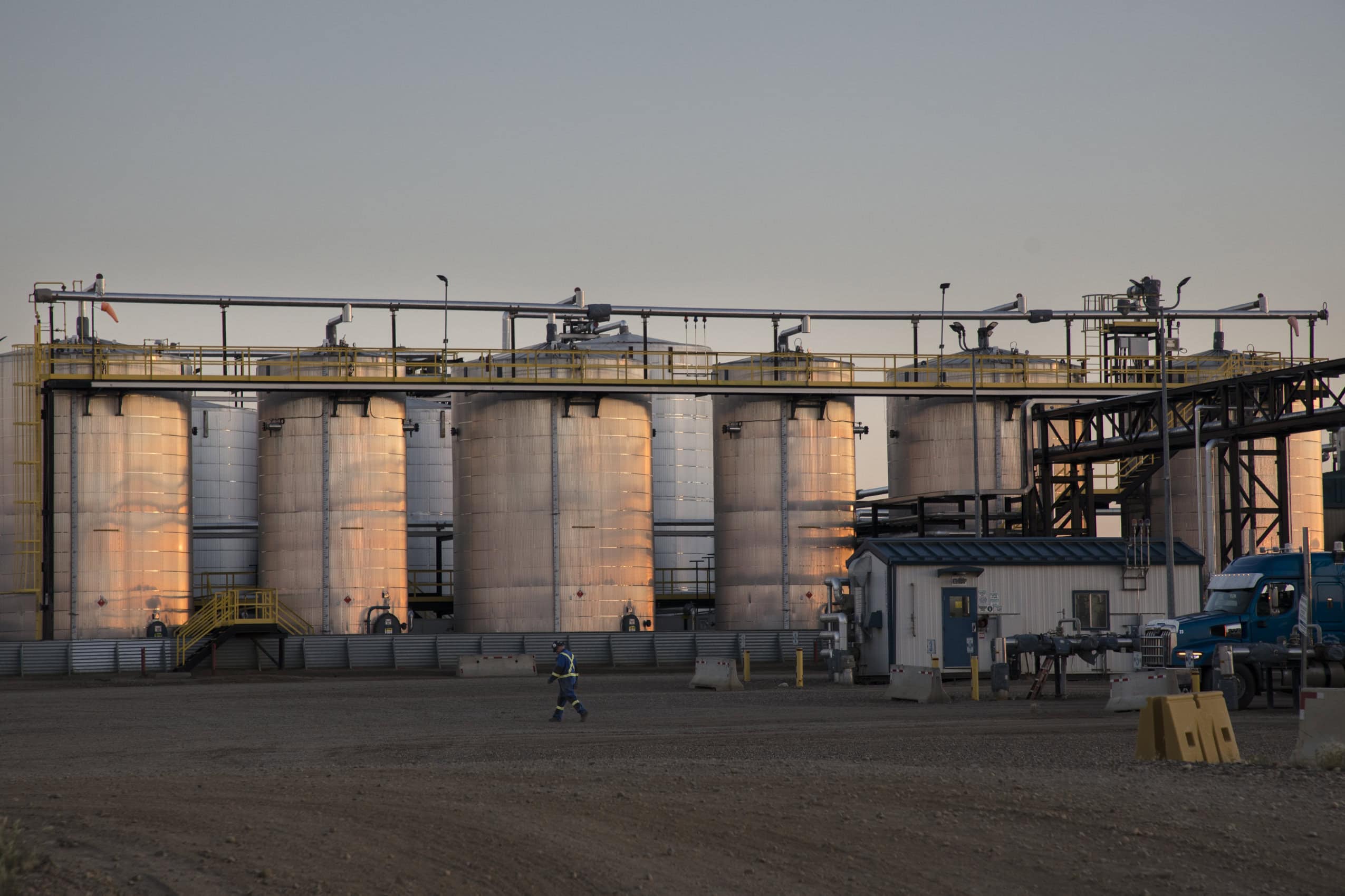 Nine large upright tanks stand along one edge of an oil and gas waste disposal facility. The sun is setting, casting a pink glow across the sides of the tanks. There's a metal walkway along with tops of the tanks. A working in a blue jump suit with reflective sites is walking across the gravel lot in front of the tanks. The blue cab of a parked heavy truck can be seen in the right corner