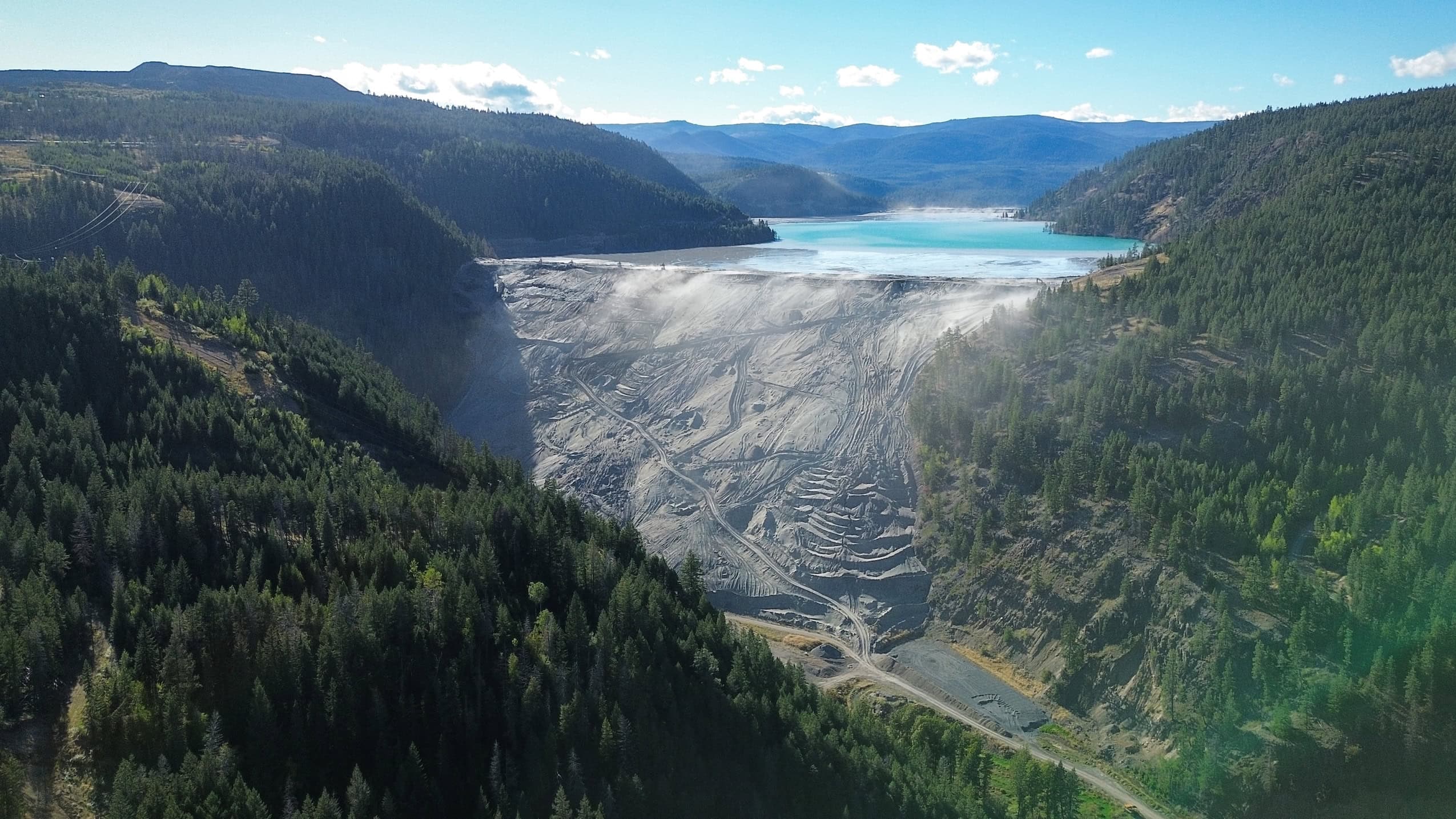 The tailings pond of a mine viewed from a distance — a bright blue pond against green forested hillsides.