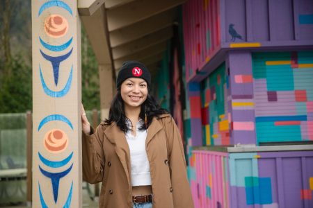 A woman wearing a brown jacket, white t-shirt and black hat with The Narwhal on it stands in front of a colourful building