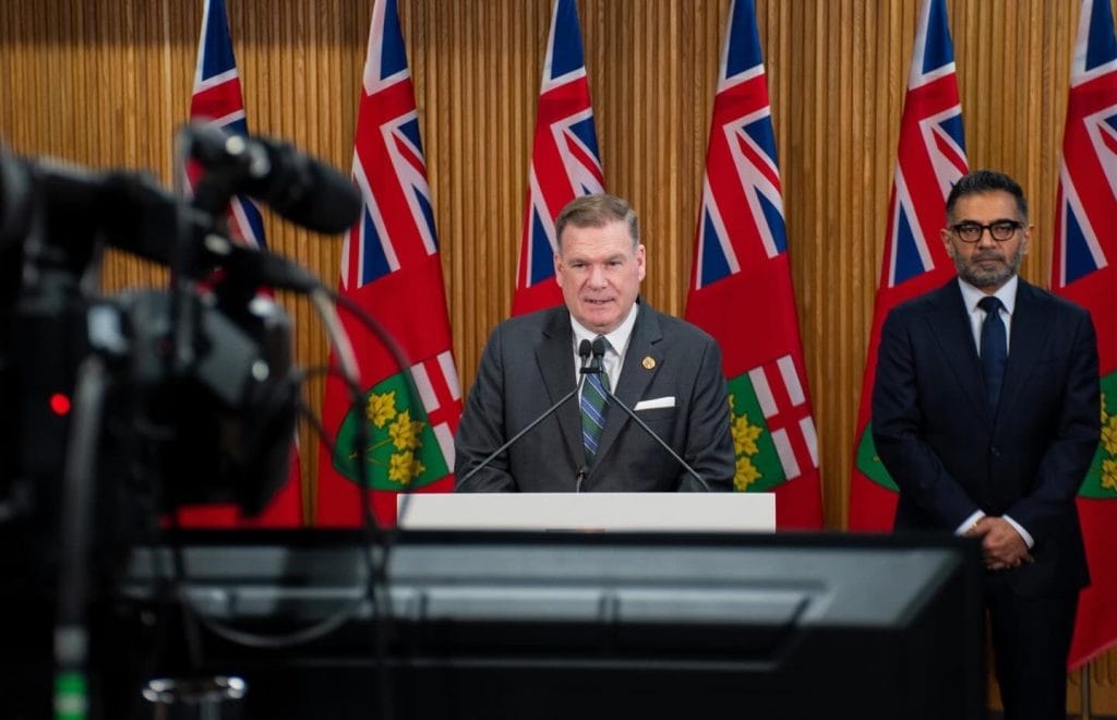 A man in a suit speaks into a microphone during a press conference, while another man stands behind him. A row of Ontario flags is in the background.