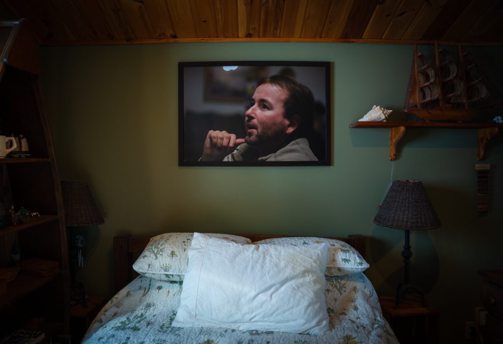 A photo of a young man hangs on a wall above a bed.