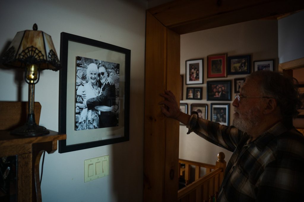 A man stands in his home and looks at a black and white photograph of him and his wife hanging on the wall.