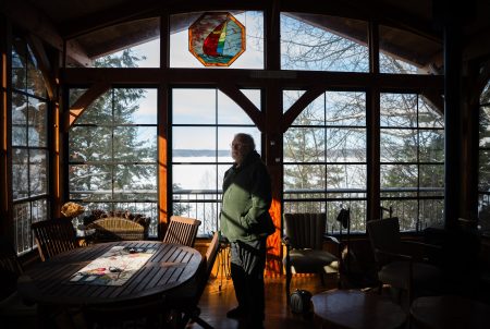 A man stands in a sun room as soft, late-day light streams in through the windows. Outside, a lake covered by ice and snow.
