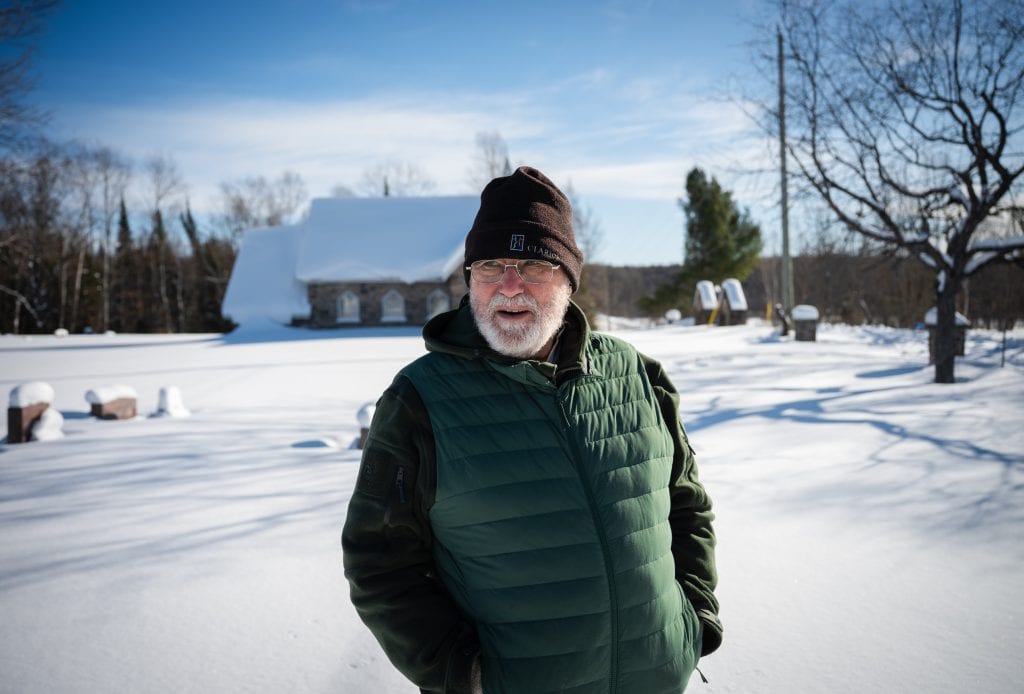 Terry Moore stands in a small cemetery in Algonquin Highlands, Ont., wearing a toque and green vest, in wintertime.