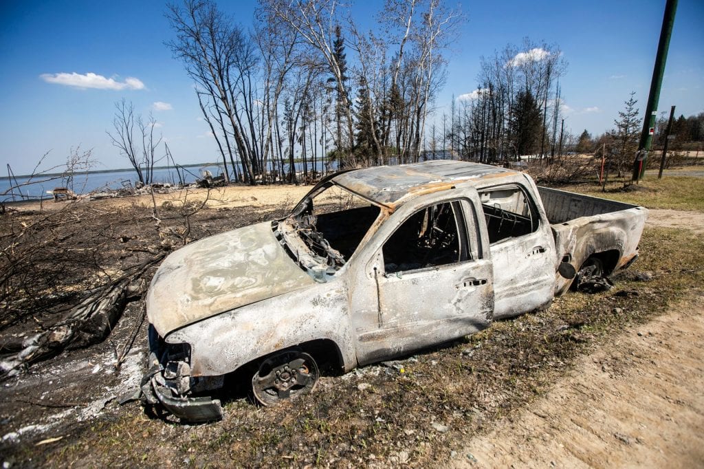 A burned-out pick-up truck and charred debris near a lakeshore.