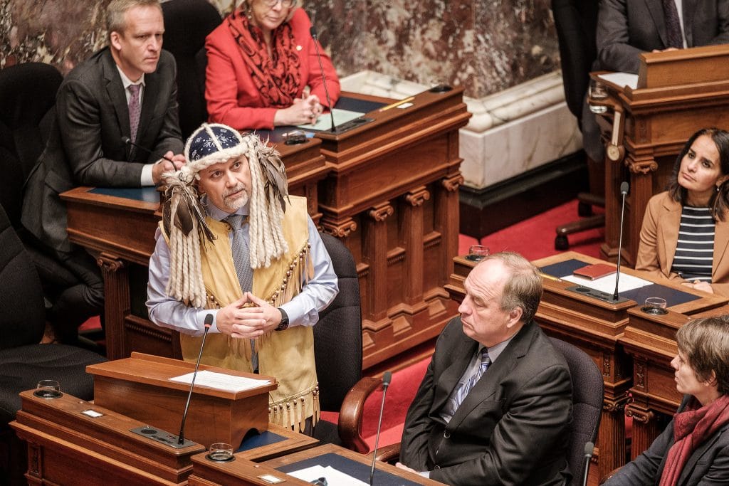 Former Green Party MLA Adam Olsen stands in the BC legislature as several MLAs look on. Olsen is wearing traditional regalia