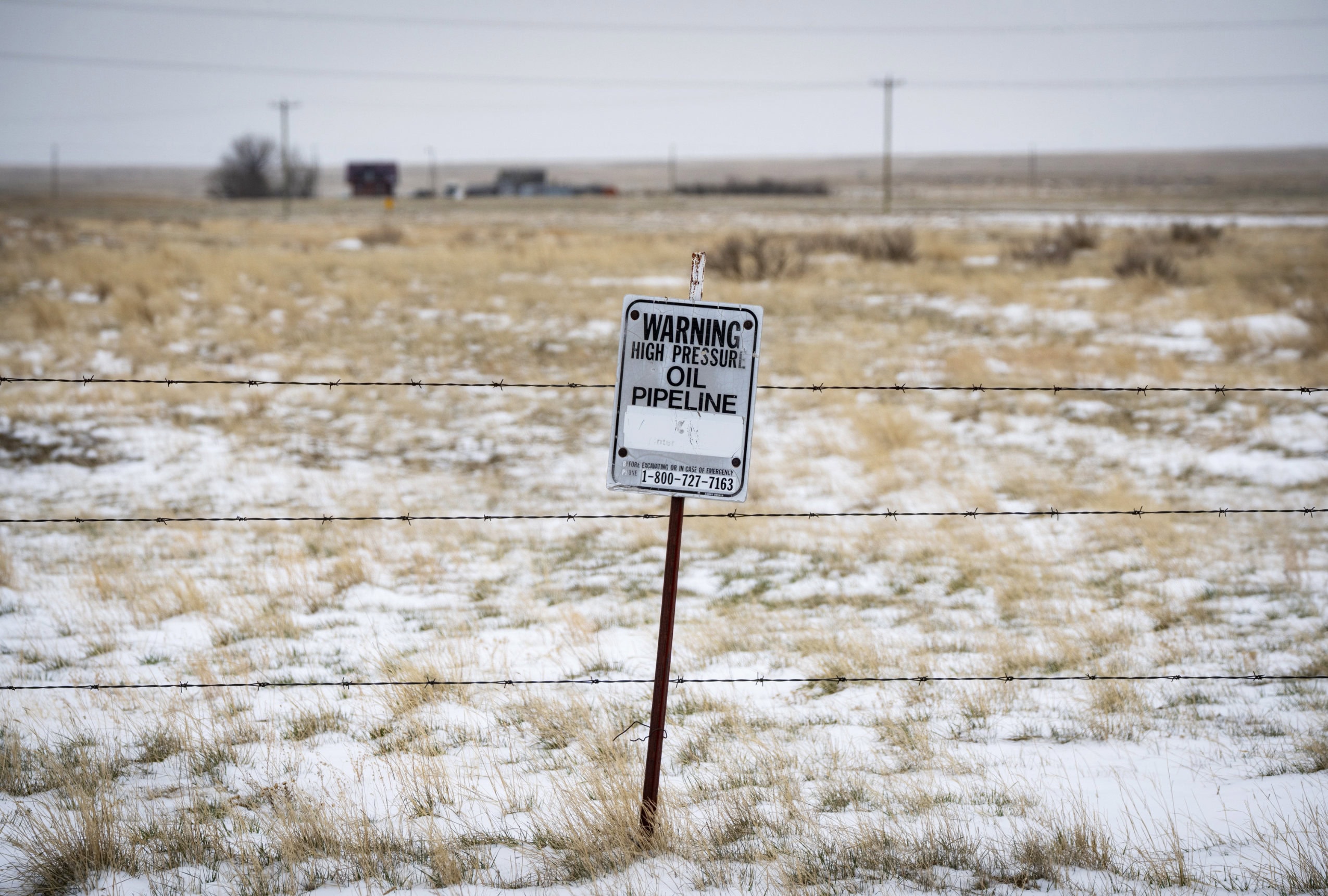 A sign reading "Warning High Pressure Oil Pipeline" stands alongside a barbed-wire fence in rural Alberta.