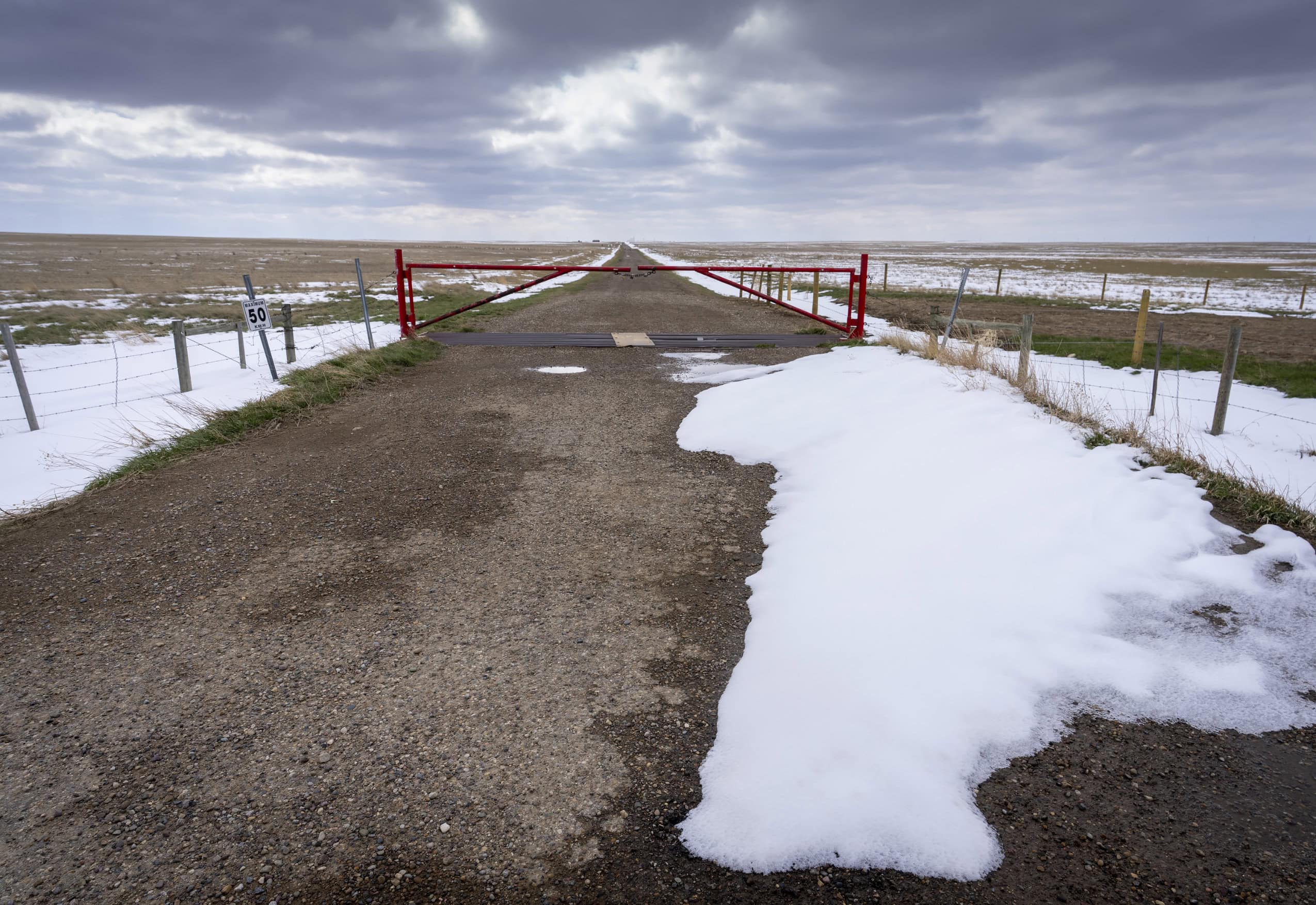 A locked gate bars entry to a road that cuts through a vast Alberta prairie landscape partially covered in snow.