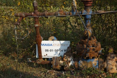 A MAGA Energy sign sits against an rusted old well site, surrounded by plants.