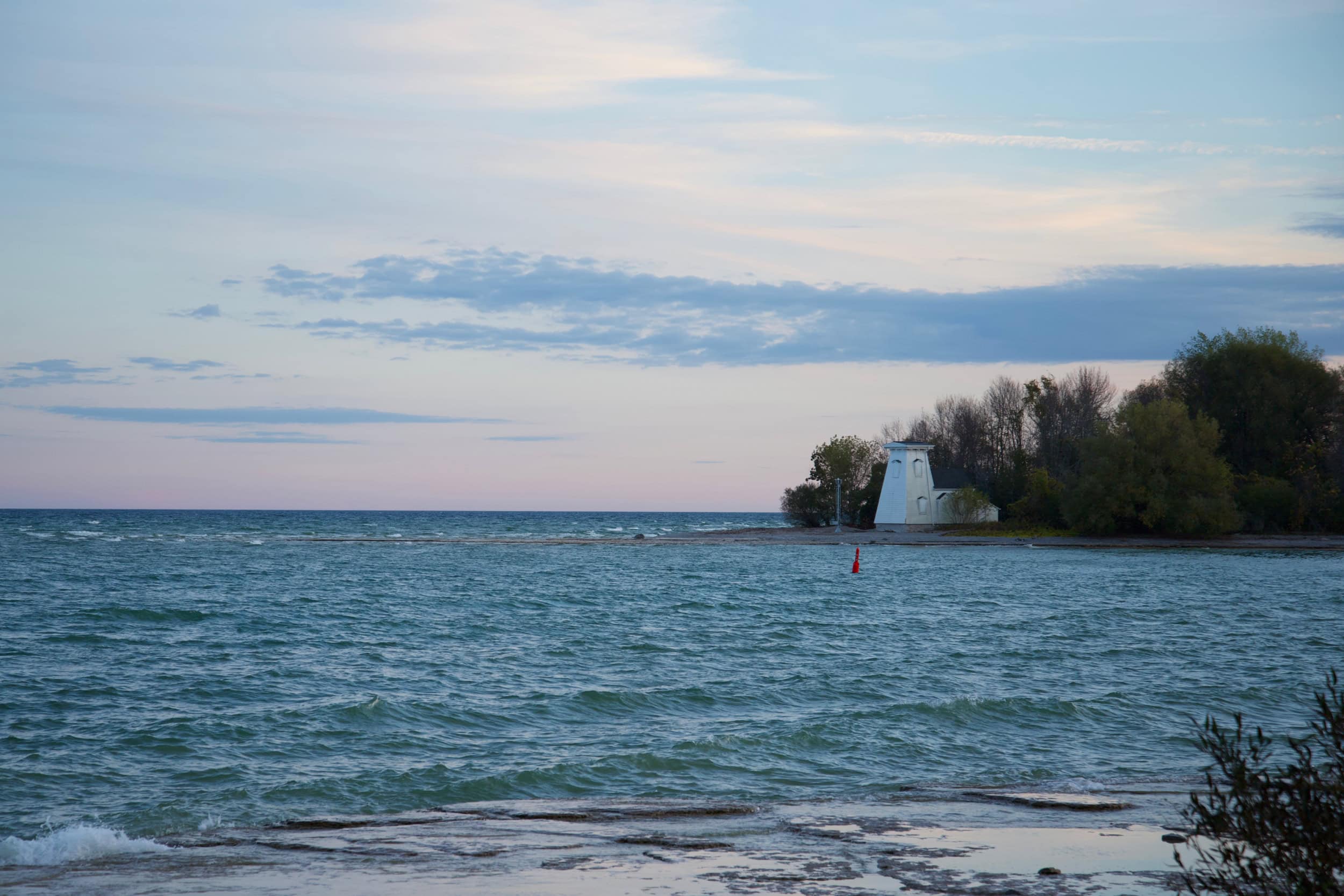 A white lighthouse on the forested point of a bay's edge, with water along the shoreline in the foreground.