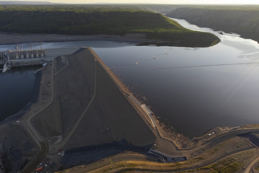 An aerial view of the Site C dam and reservoir at dusk. Looking across the Peace River, which the dam spans, there are green hills and hazy summer sky in the background