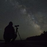 A stargazer with a camera is silhouetted against the milky way in the night sky.