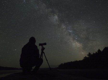 A stargazer with a camera is silhouetted against the milky way in the night sky.