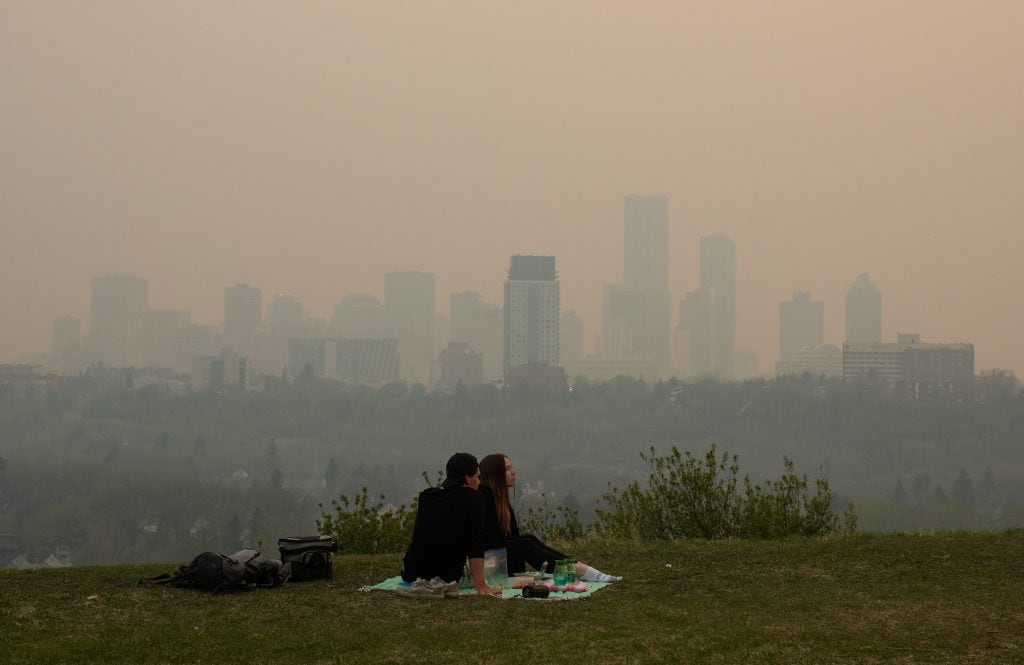 Two people sit on a picnic blanket as smoke hangs over the Edmonton skyline in the background.