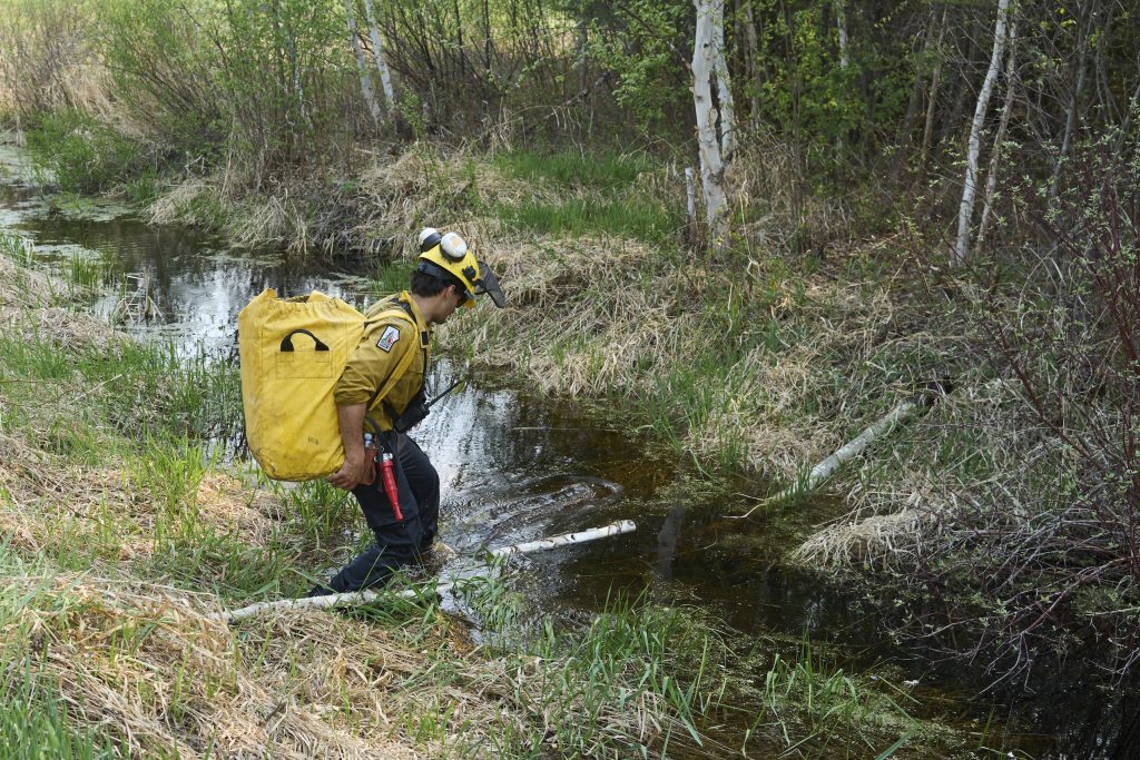 A wildfire fighter crosses a stream with a hose on his back.