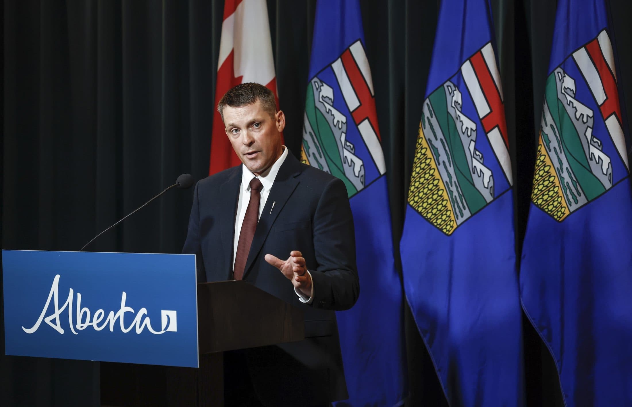 Alberta Finance Minister speaks at a lectern during a news conference, with Canadian and Albertan flags behind him.