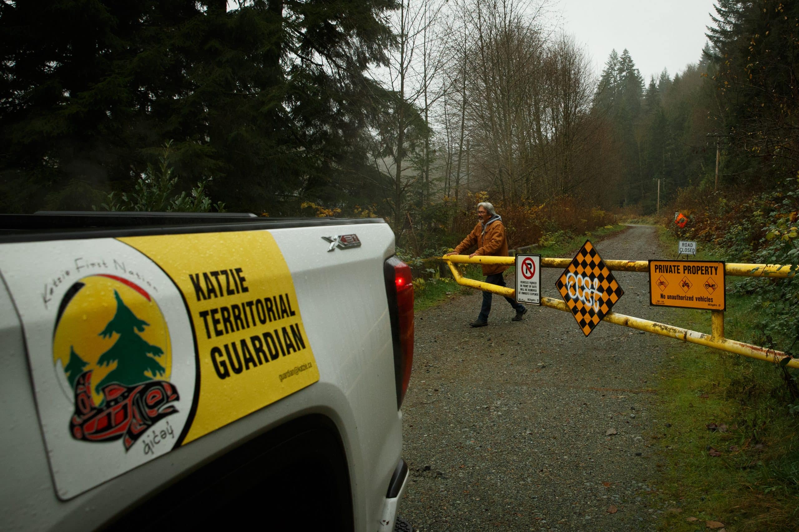 A man closing a gate that leads into a trail in the forest, with a white truck in front that says "Katzie territorial guardian" on it.