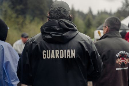 The back of a man's jacket has the word guardian printed on it with white lettering as he looks into a crowd.