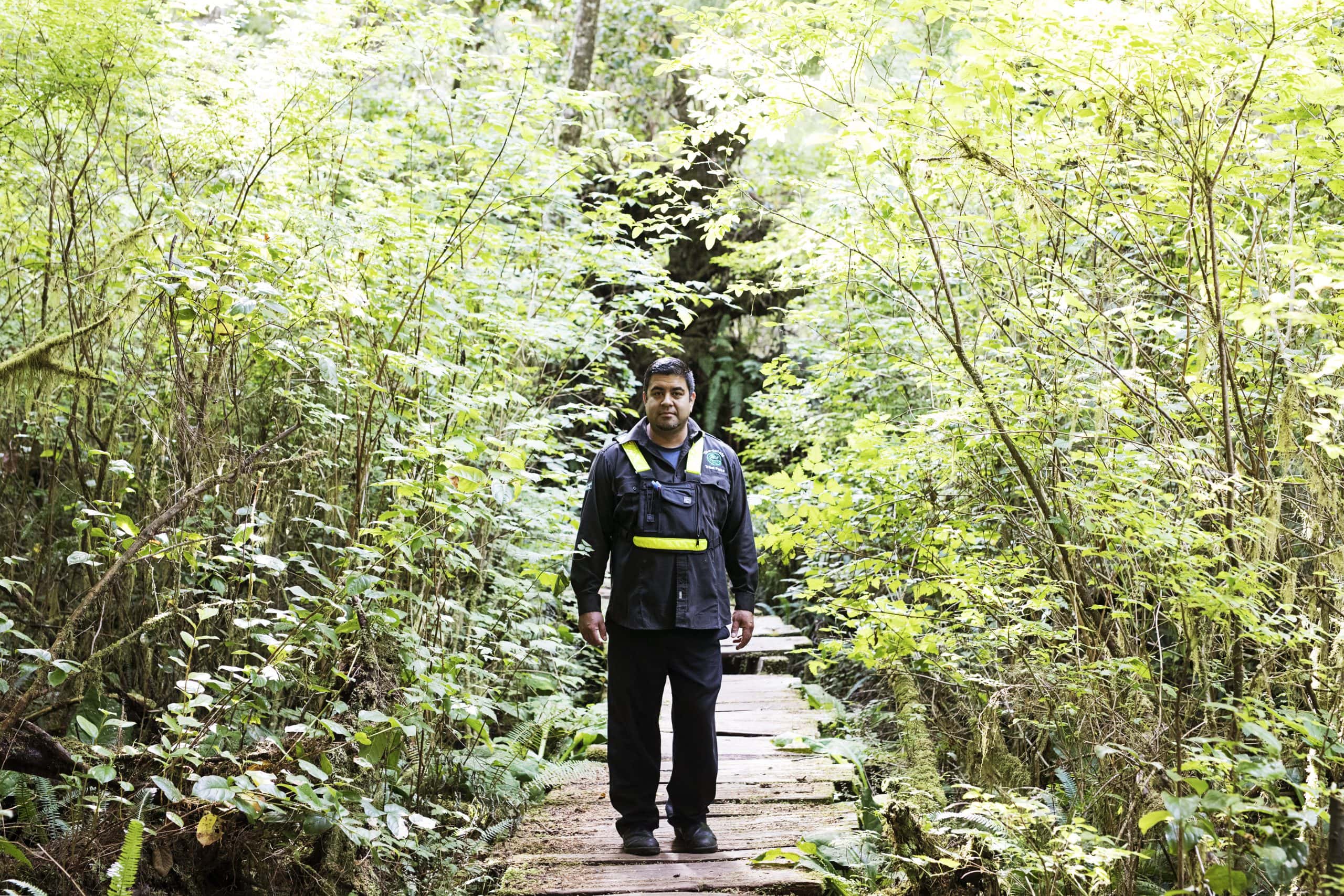 A man wearing all black stands in a walking trail with lush greenery around him.