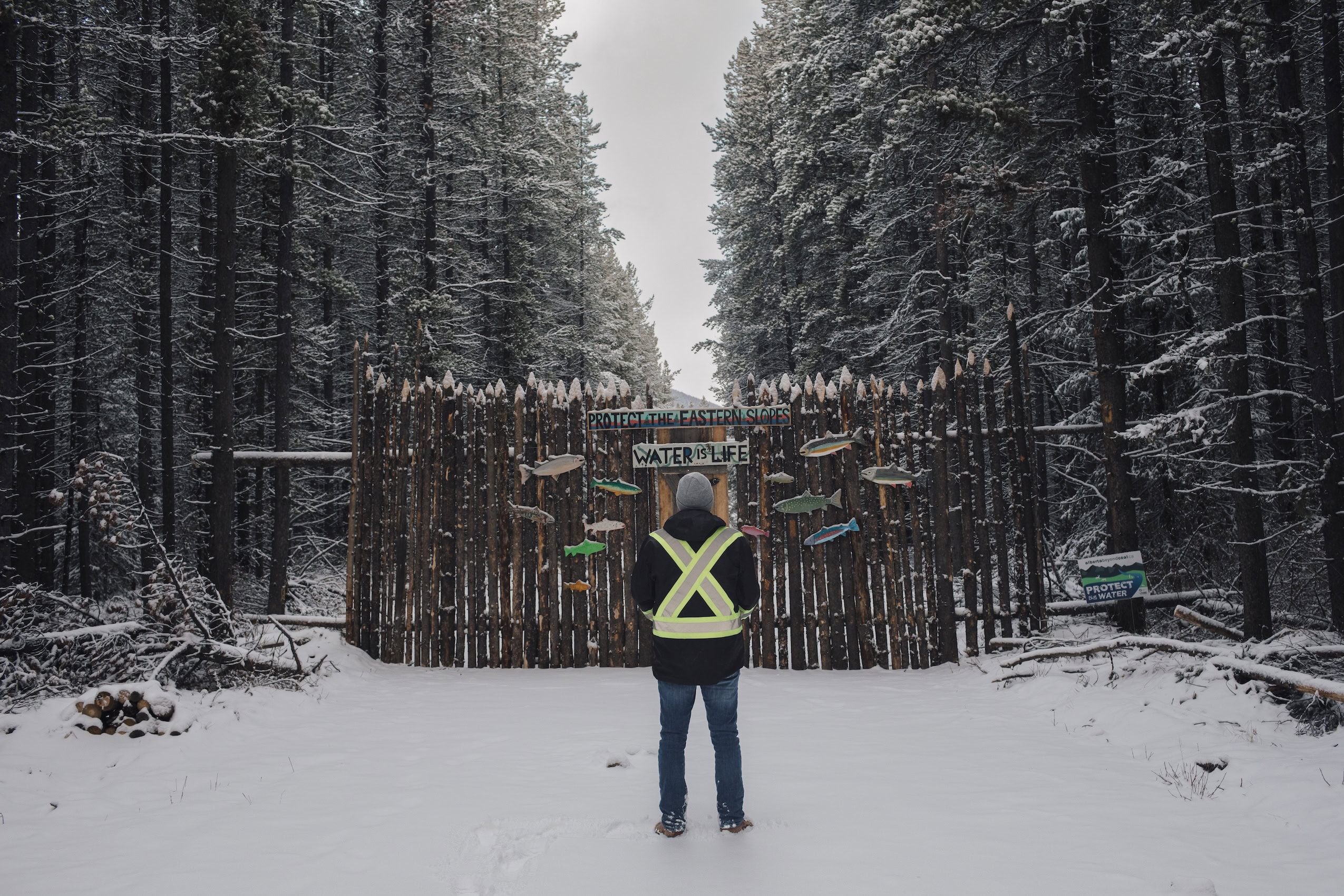 A man with a reflective vest stands with his back to the camera, in front of a log fence with the words "water is life" on it.