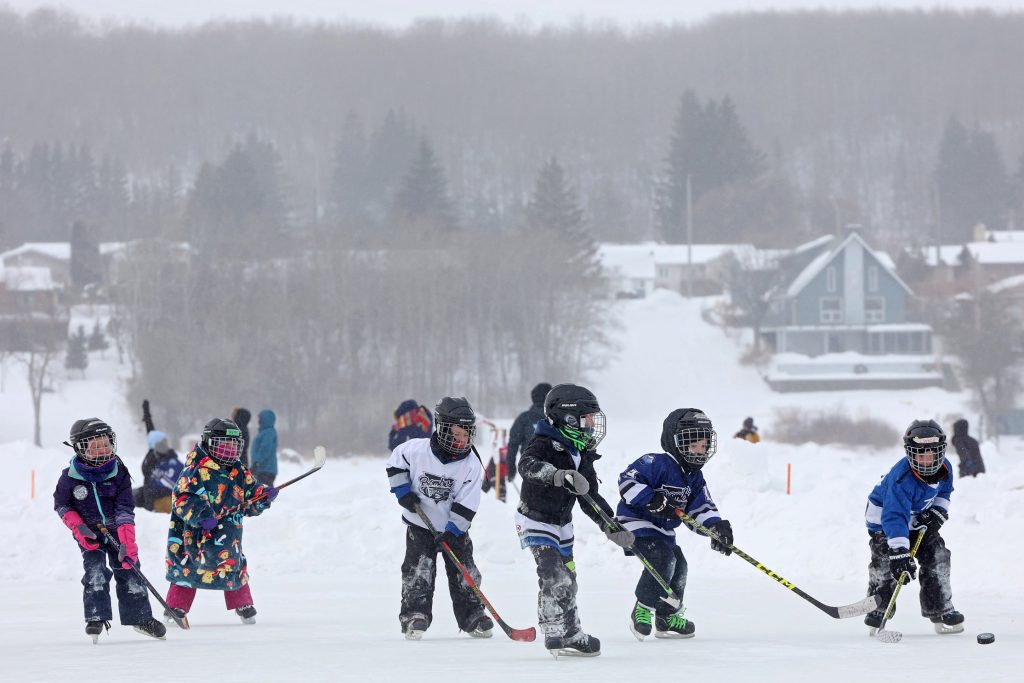 Young children in hockey jerseys and warm winter gear play hockey on the ice on a wintry day