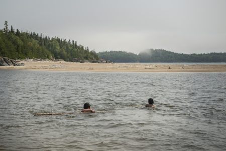 Two people swim in Lake Superior, with a sandy shoreline in the background.