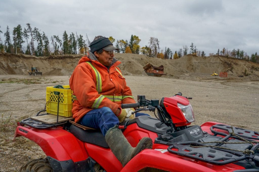 A man wearing an orange safety coat with reflective yellow elements sits on a fourwheeler, with a gravel pit in the background.