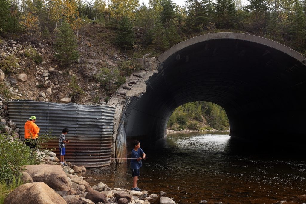 A man and two children fish on a rocky bank of a river near a large culvert.