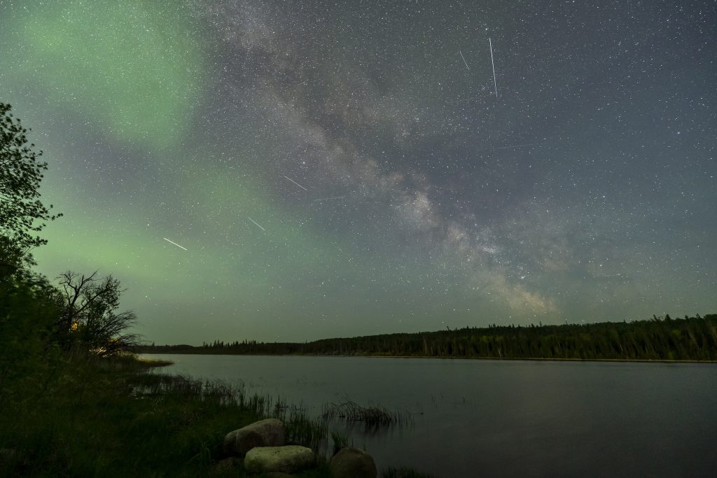 The northern lights and stars light up the night sky, with a quiet lake in the foreground.