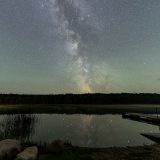 The northern lights and stars light up the night sky, with a quiet lake in the foreground.
