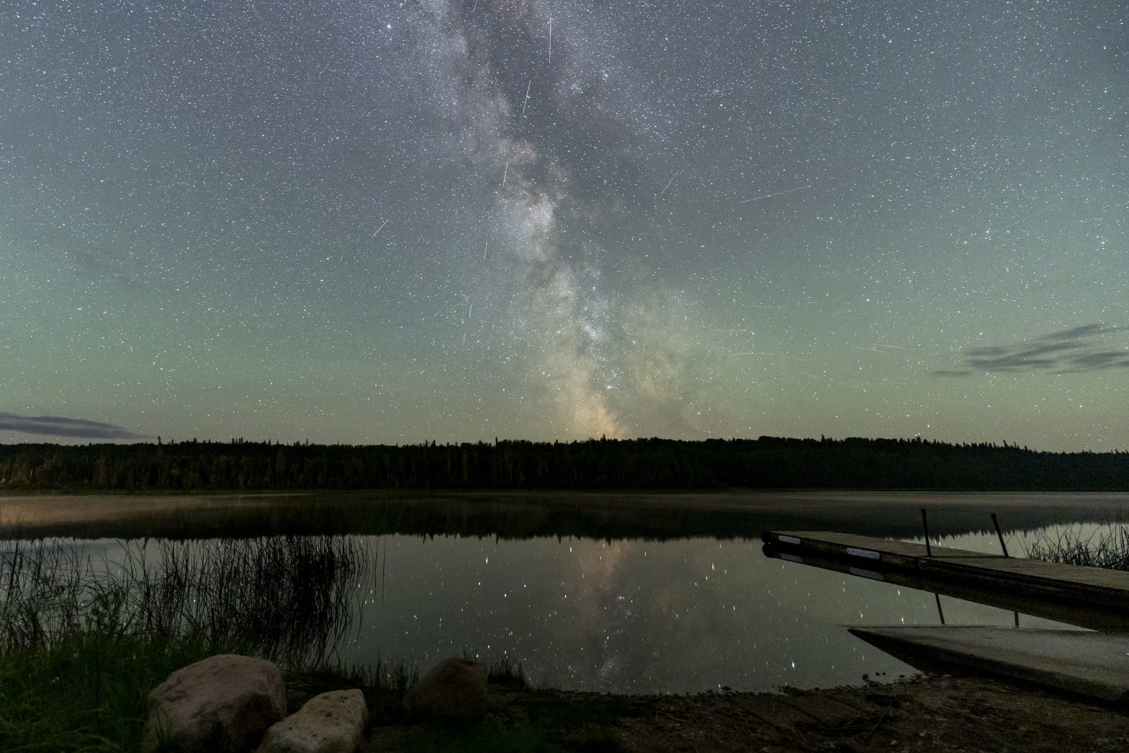 The northern lights and stars light up the night sky, with a quiet lake in the foreground.