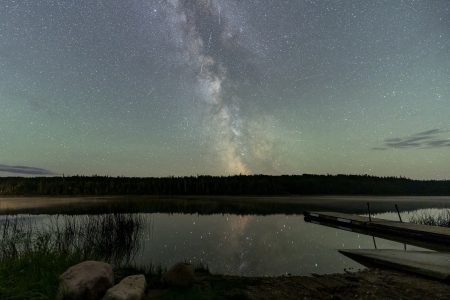 The northern lights and stars light up the night sky, with a quiet lake in the foreground.