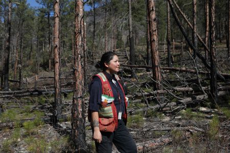 A woman wearing a work vest looks off into the distance in front of trees.