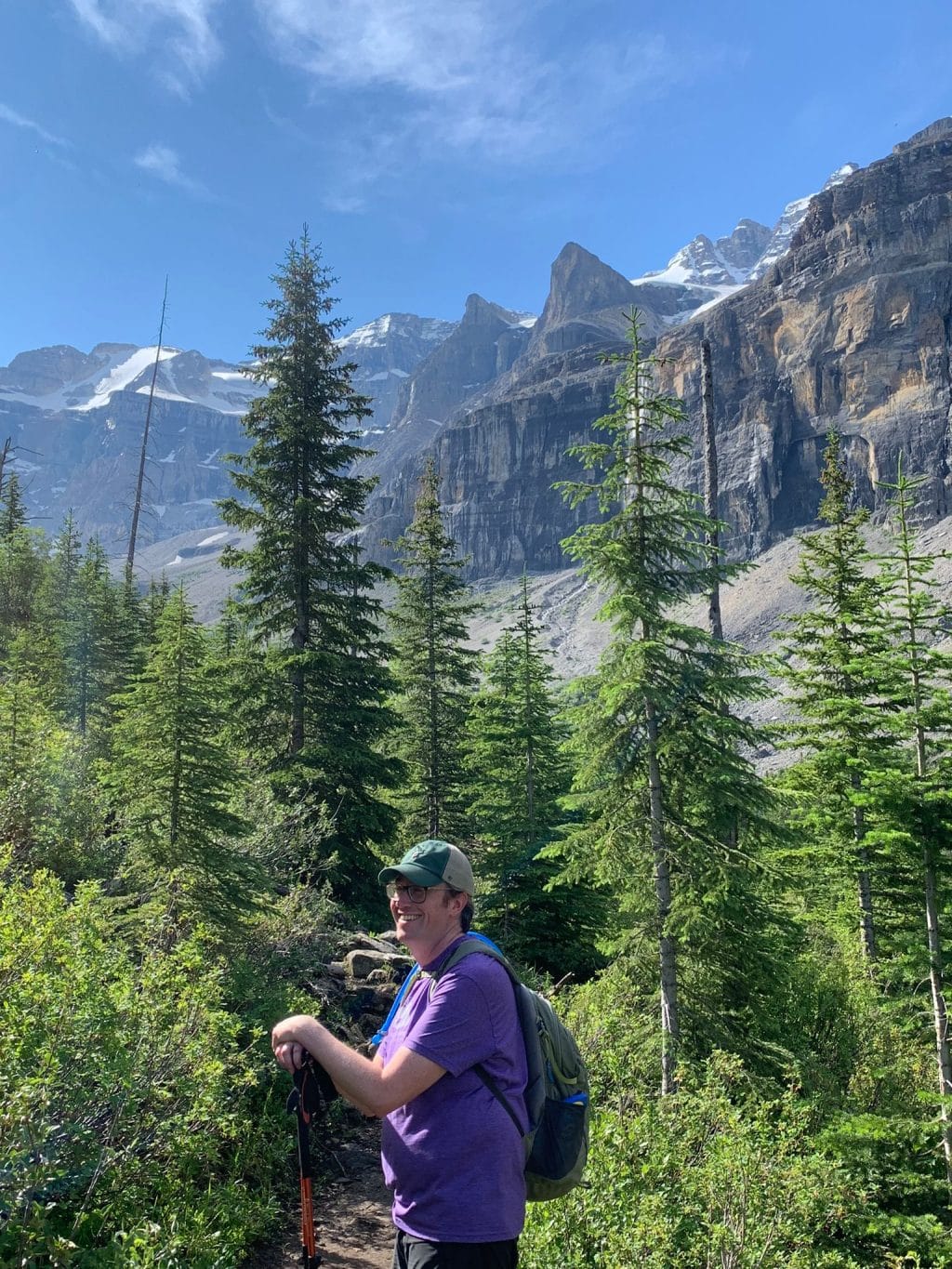 Alex Goatcher stands outdoors with hiking gear. Trees and mountains are behind him.