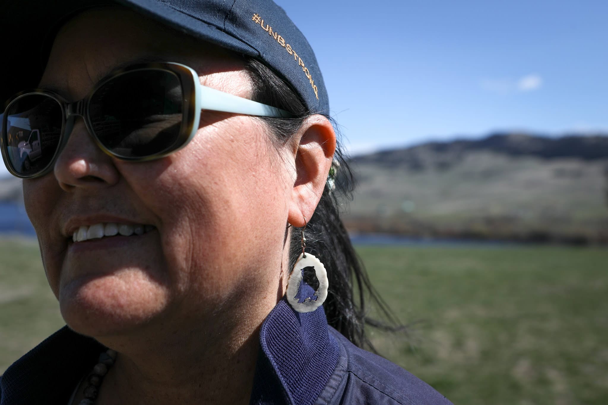 A woman in sunglasses and a blue hat wearing owl earrings smiles