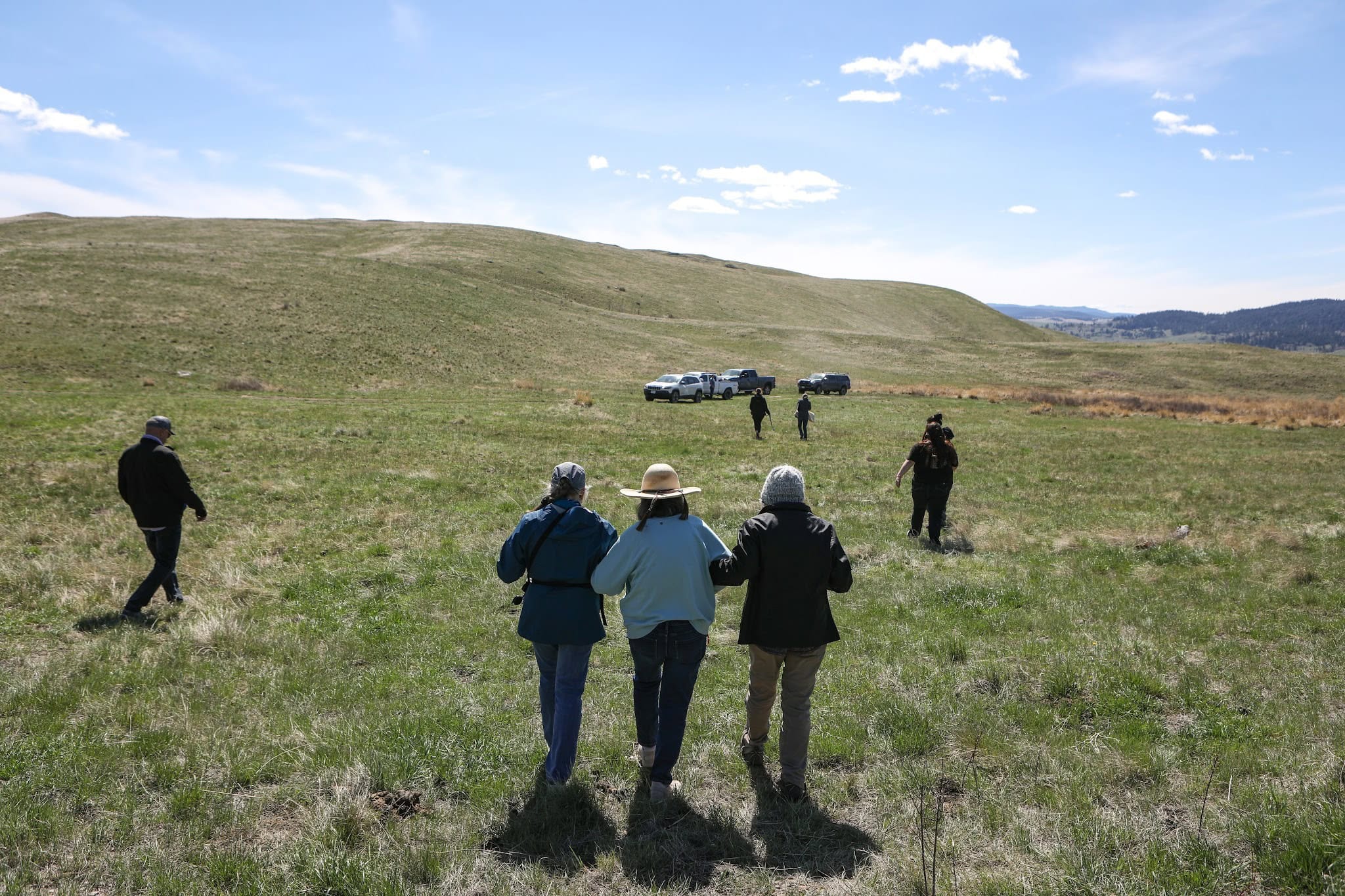 Three community members walk across a grassy field toward a hill, with trucks parked in the distance. 