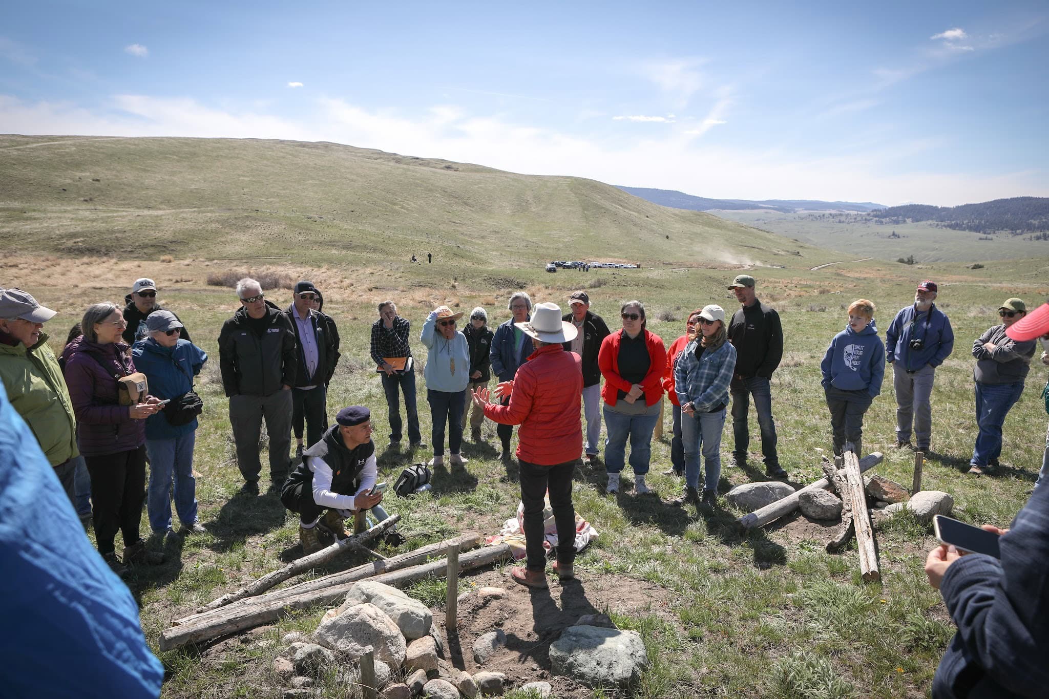 A woman in a red jacket and light cowboy hat lectures to an assembled crowd in a grassy field during an owl release.