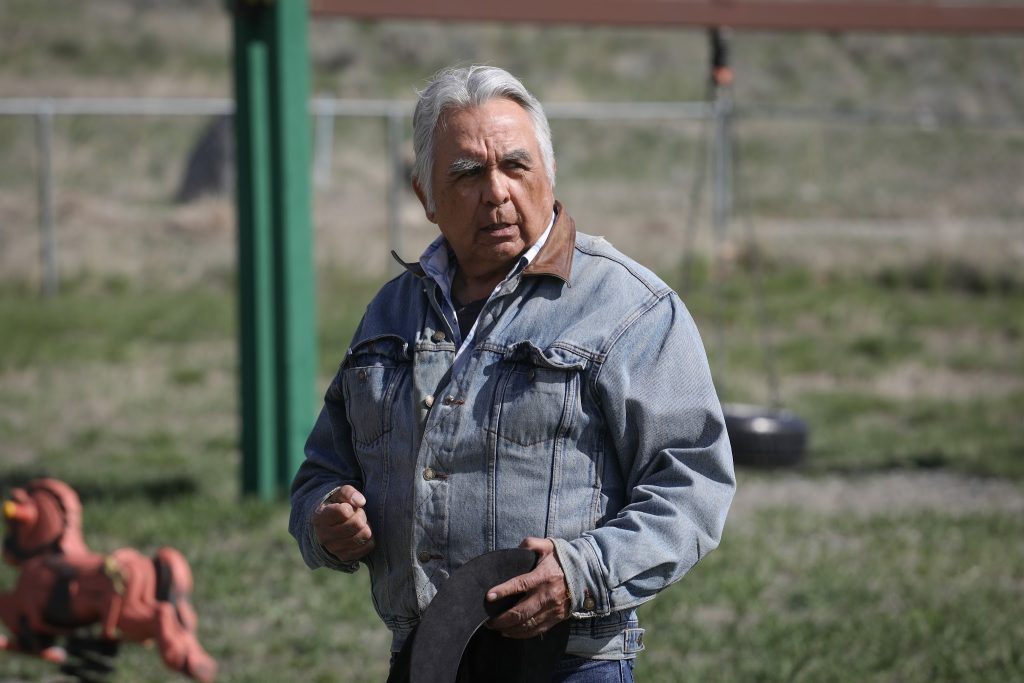 An older man with white hair and a denim jacket speaks in front of a playground.