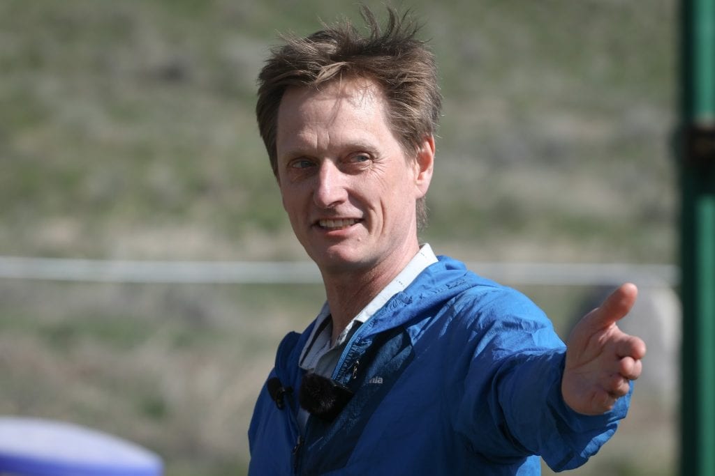 A man with brown hair in a blue windbreaker gestures toward the camera. 